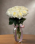 Bouquet of white roses in a clear vase with a pink ribbon on a wooden surface.