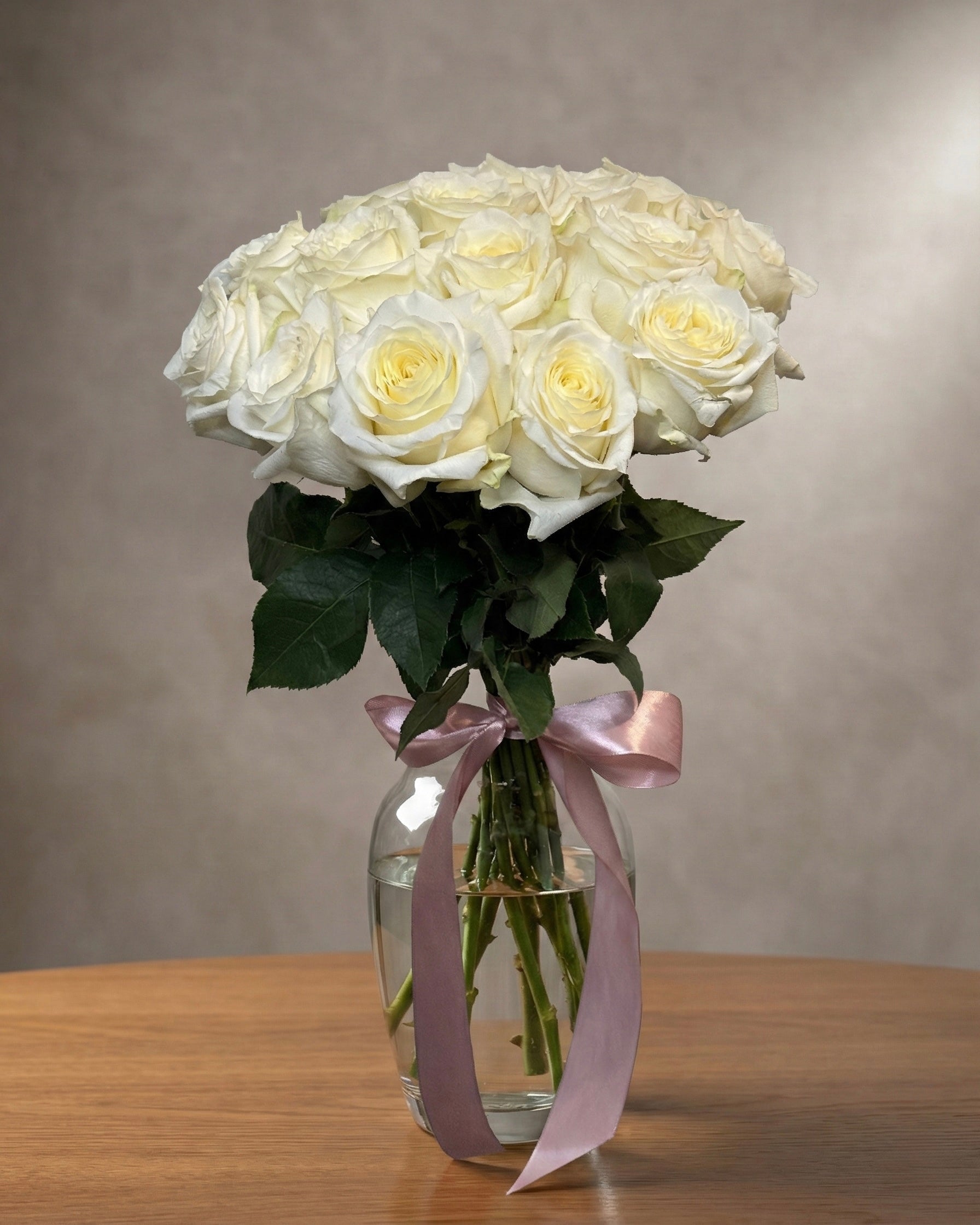 Bouquet of white roses in a clear vase with a pink ribbon on a wooden surface.
